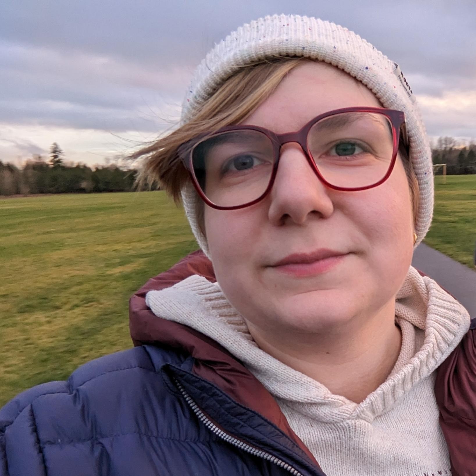 A photo of Alex wearing a white beanie and a hooded sweatshirt layered under a down jacket. They are standing in a field.